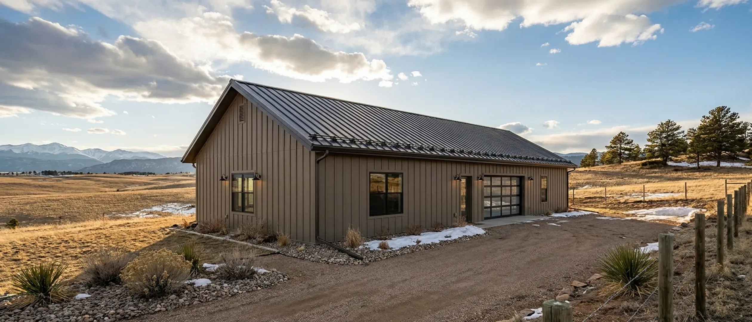 A completed barndominium viewed from a low angle showing its standing seam metal roof and vertical wall panels, with the last patches of snow melting beside it.